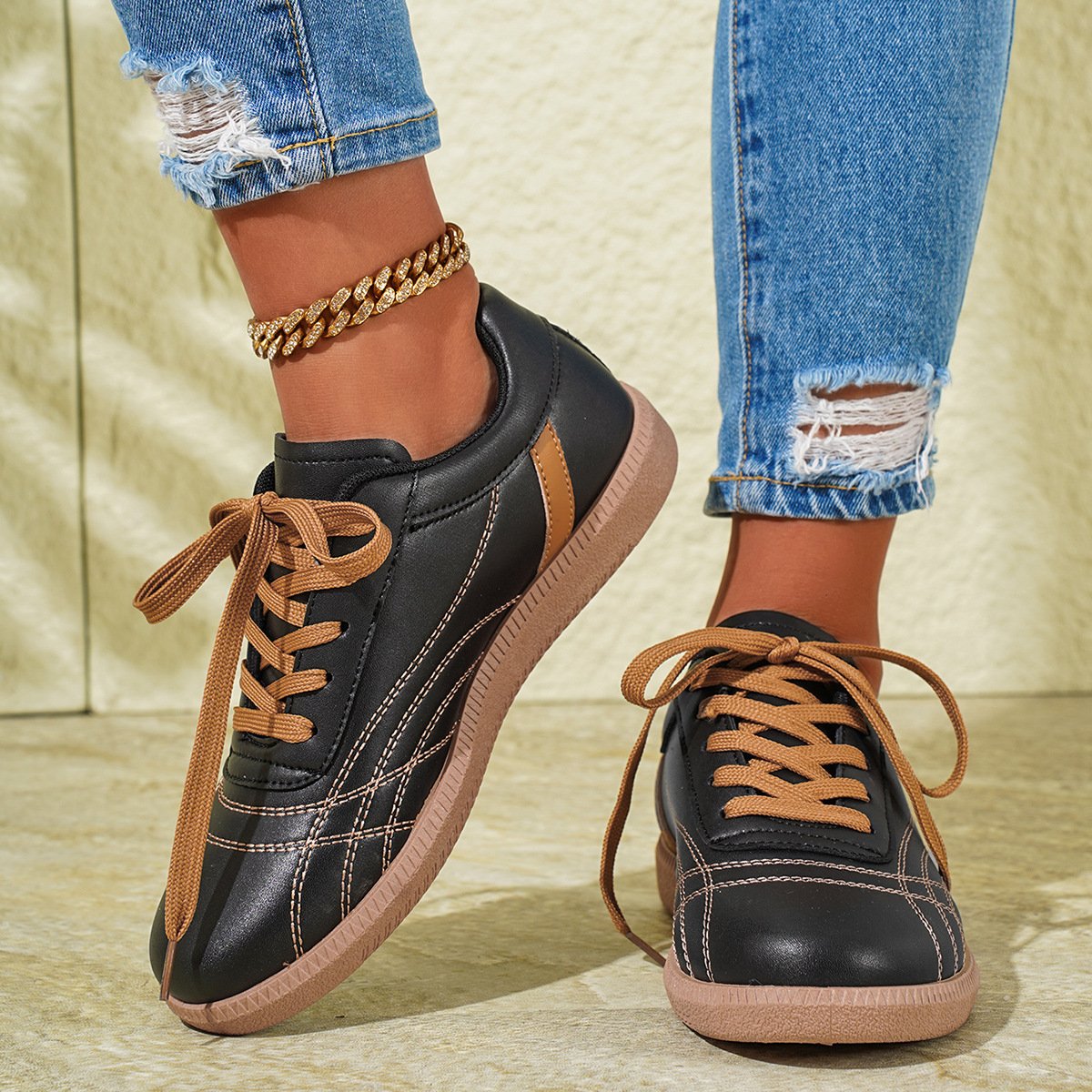 Damen Sneaker in Schwarz mit braunen Schnürsenkeln, kombiniert mit zerrissenen Jeans und goldener Fußkette. Modische Freizeitschuhe für den Alltag.