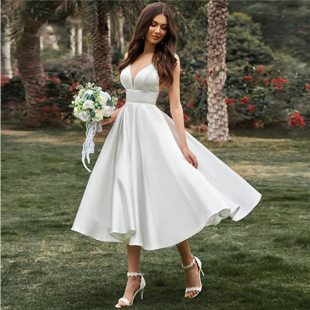 Braut in weißem Hochzeitskleid mit Blumenstrauß im Garten, umgeben von Palmen und Rosen. Elegantes, ärmelloses Design, ideal für Sommerhochzeiten.
