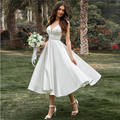 Braut in weißem Hochzeitskleid mit Blumenstrauß im Garten, umgeben von Palmen und Rosen. Elegantes, ärmelloses Design, ideal für Sommerhochzeiten.