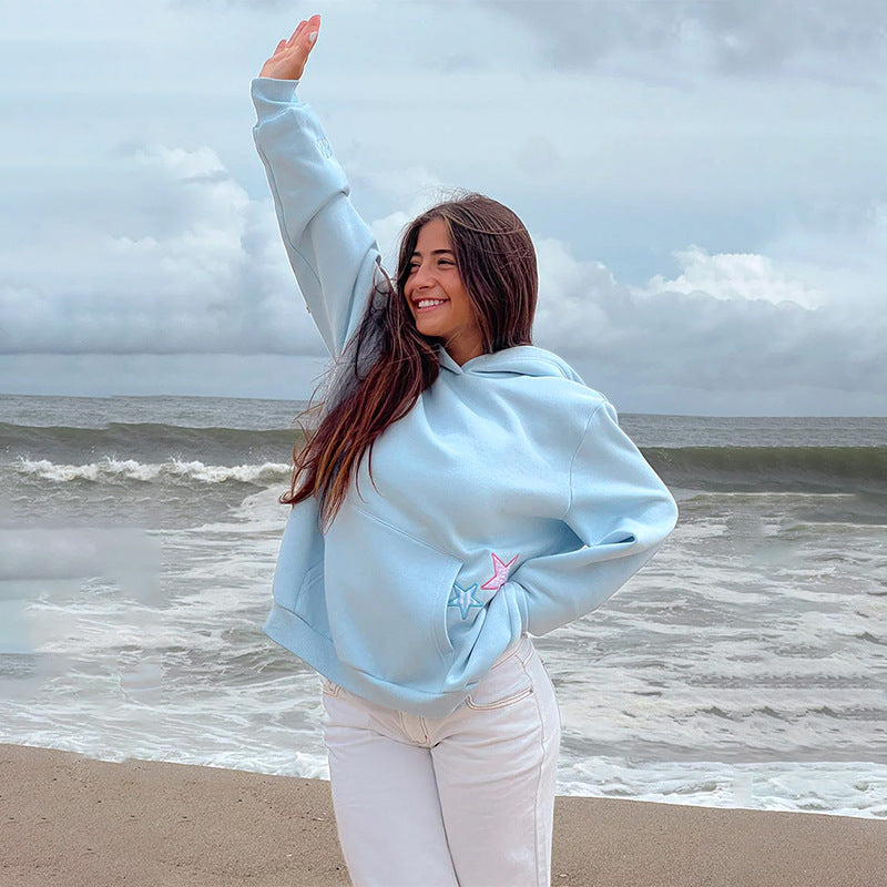 Frau am Strand in hellblauem Hoodie, lächelnd und winkend vor stürmischer See. Freizeitmode, Strandspaziergang, Herbstwetter.