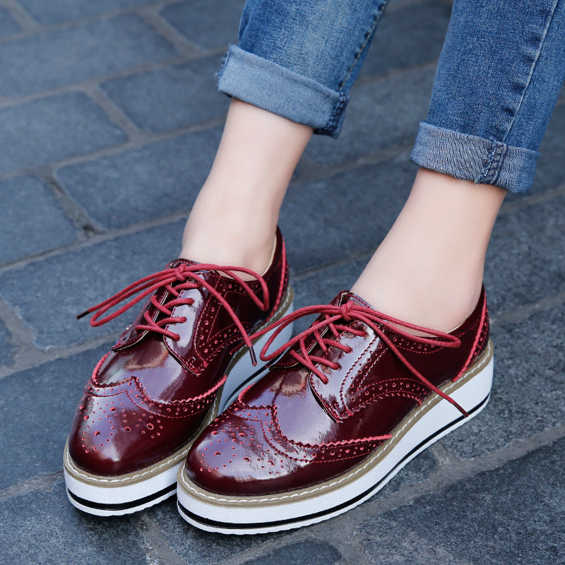 Damenfüße in blauen Jeans tragen glänzende, rote Brogues mit Plateauabsatz auf gepflastertem Gehweg. Modische Schuhe, Streetstyle, Herbstmode.