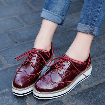 Damenfüße in blauen Jeans tragen glänzende, rote Brogues mit Plateauabsatz auf gepflastertem Gehweg. Modische Schuhe, Streetstyle, Herbstmode.