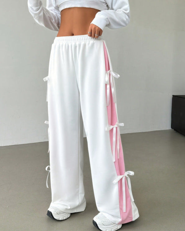 Frau in weißer Jogginghose mit rosa Streifen und Schleifen, bauchfreies Oberteil, modische Sportbekleidung, trendige Damenmode, Fitness-Outfit.