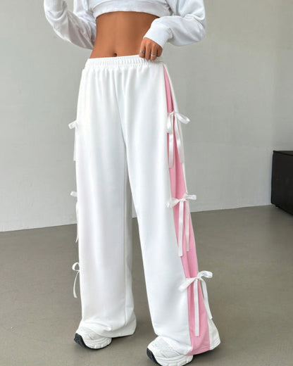 Frau in weißer Jogginghose mit rosa Streifen und Schleifen, bauchfreies Oberteil, modische Sportbekleidung, trendige Damenmode, Fitness-Outfit.