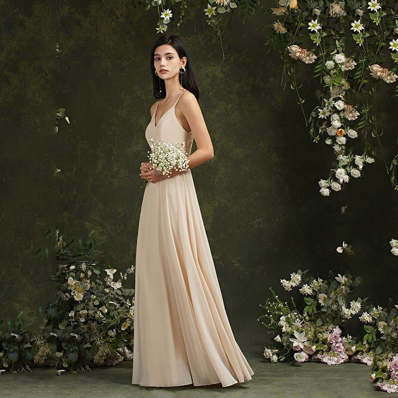 Elegante Frau im langen, cremefarbenen Abendkleid mit Blumenstrauß, umgeben von blühenden Pflanzen vor grünem Hintergrund. Ideal für Hochzeitsmode.