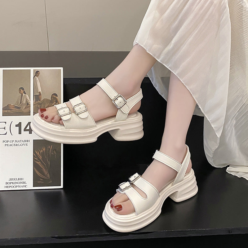 Weiße Plateau-Sandalen mit Schnallen an den Füßen einer Person in einem fließenden Kleid, neben einem Modebuch auf einem schwarzen Tisch.