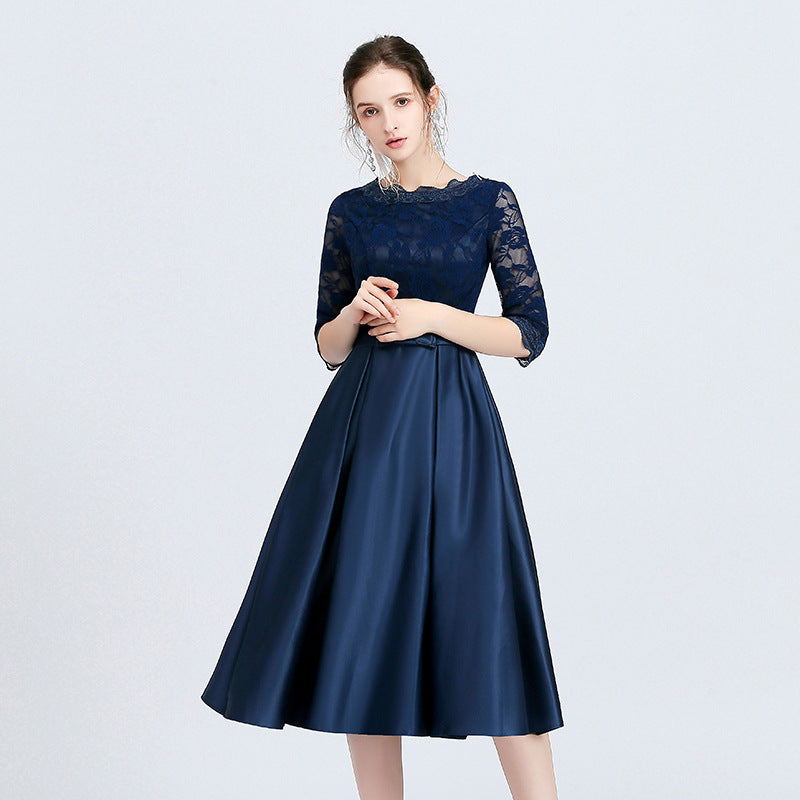 Frau in elegantem, dunkelblauem Abendkleid mit Spitzenoberteil und halblangen Ärmeln, stehend vor hellem Hintergrund.