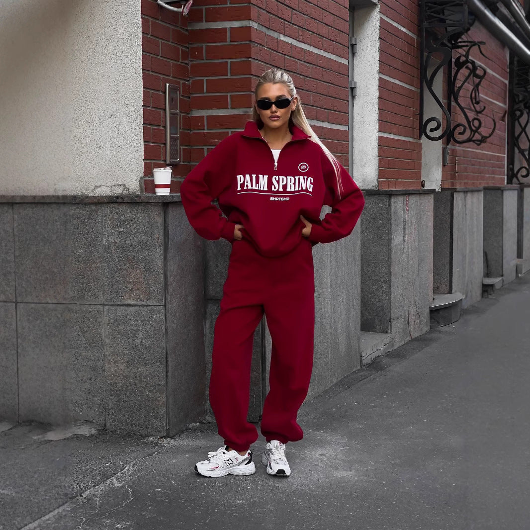 Frau in rotem Trainingsanzug mit "Palm Spring" Aufdruck, trägt Sonnenbrille und weiße Sneakers, steht vor Backsteinwand. Streetwear, urbaner Stil.