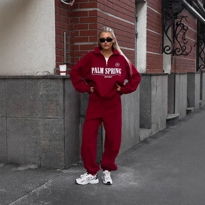 Frau in rotem Trainingsanzug mit "Palm Spring" Aufdruck, trägt Sonnenbrille und weiße Sneakers, steht vor Backsteinwand. Streetwear, urbaner Stil.