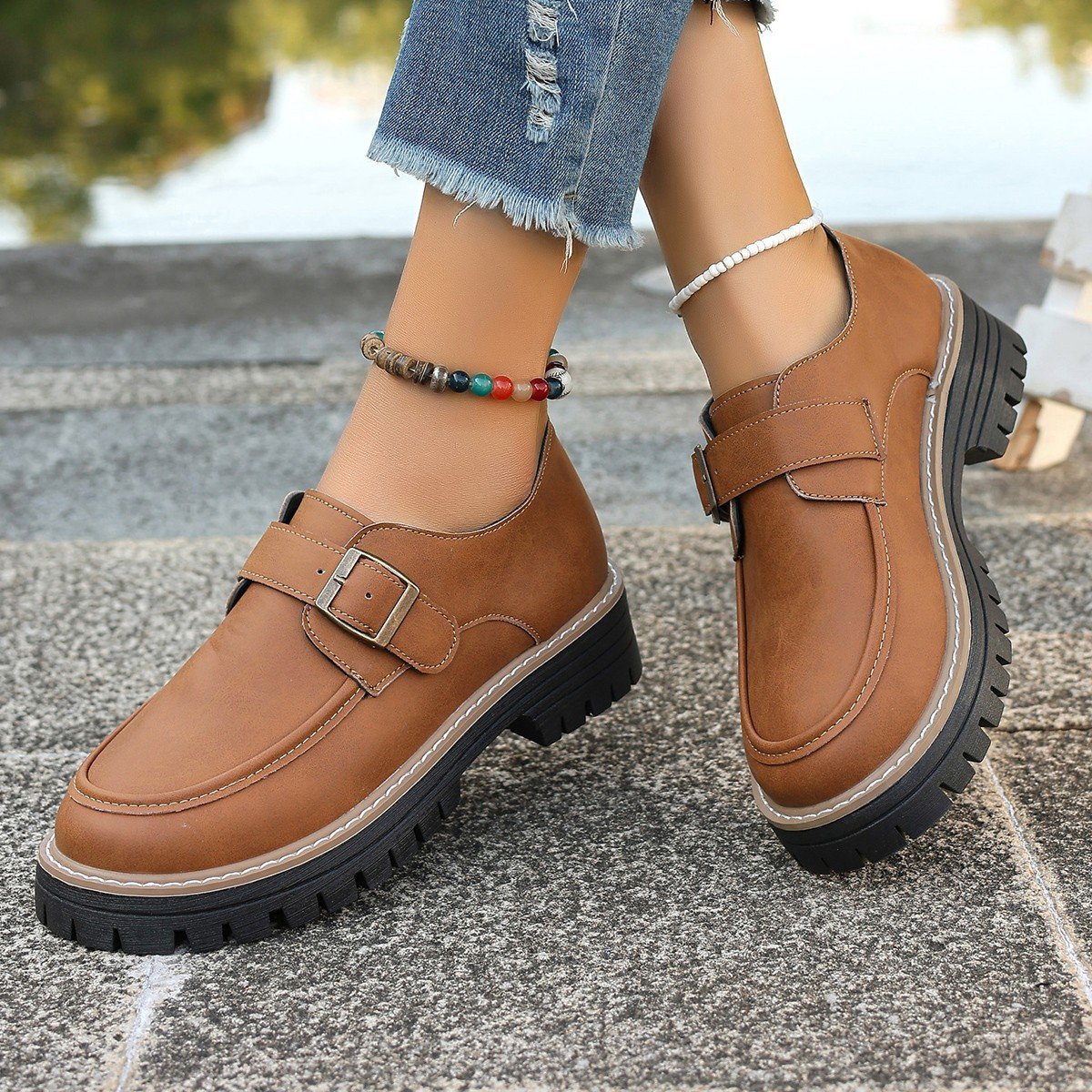 Braune Damen-Loafer mit Schnalle, robuste Profilsohle, getragen mit Jeans und buntem Fußkettchen. Modische Herbstschuhe, Outdoor-Stil.