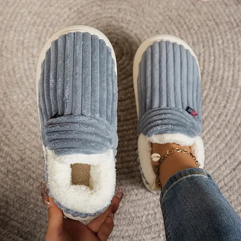 Graue, plüschige Hausschuhe mit Rillenmuster auf einem Teppich, getragen von einer Person mit Jeans. Bequeme, warme Pantoffeln für Zuhause.