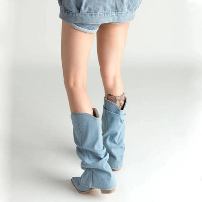 Frau in blauen Jeans-Shorts und hellblauen Cowboy-Stiefeln mit Tattoo am Bein. Modetrend, Denim-Look, Sommeroutfit, Streetstyle.
