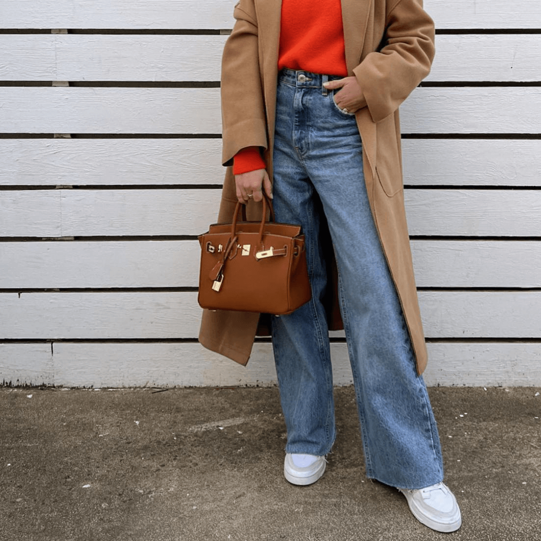 Frau in beigem Mantel und rotem Pullover, trägt blaue Jeans und weiße Sneaker, hält braune Designer-Handtasche vor weißem Holzzaun.