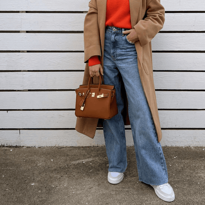 Frau in beigem Mantel und rotem Pullover, trägt blaue Jeans und weiße Sneaker, hält braune Designer-Handtasche vor weißem Holzzaun.