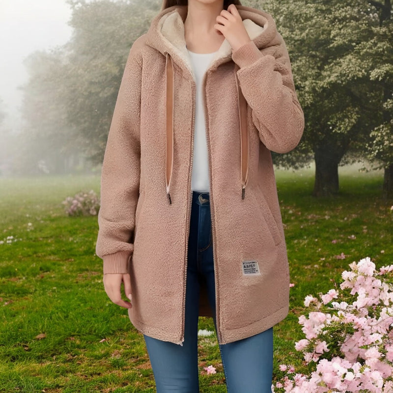 Frau in beigem Teddy-Mantel im Park, umgeben von blühenden Bäumen und Blumen. Modetrend Herbst, warme Damenbekleidung, Outdoor-Mode.