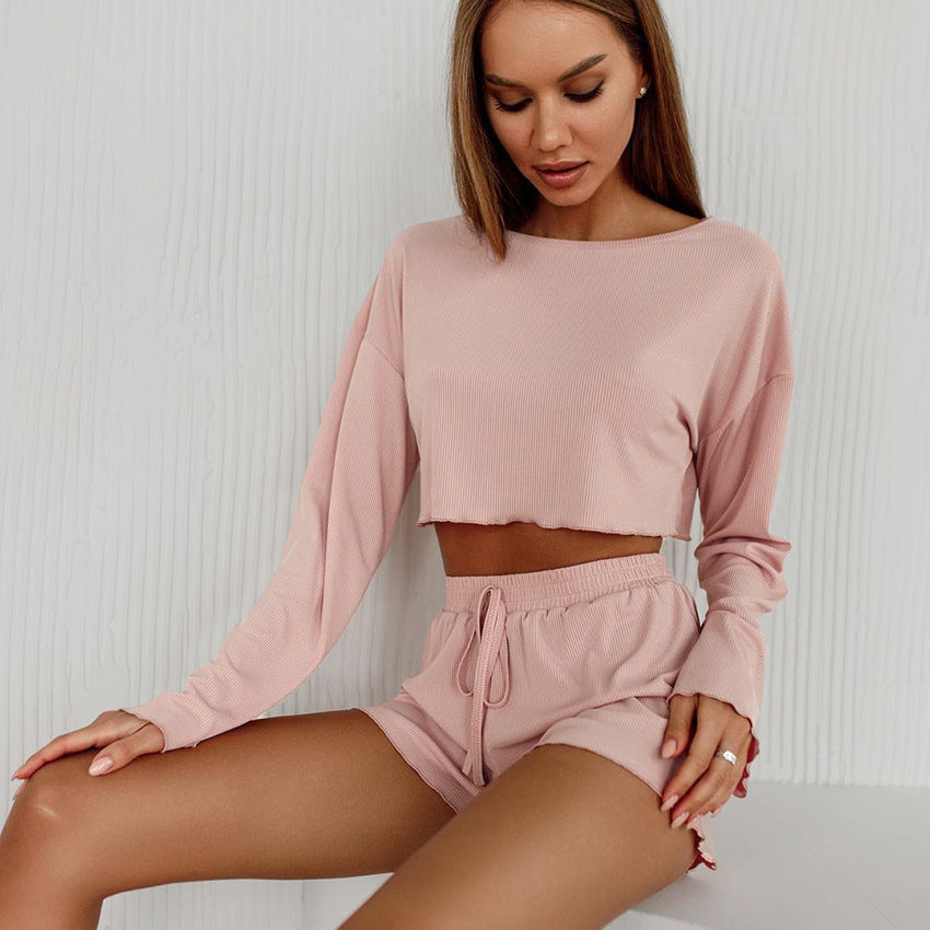 Frau in rosa Lounge-Set, bestehend aus langärmligem Crop-Top und Shorts, sitzt vor weißem Hintergrund. Bequeme Damenmode, Freizeitkleidung.