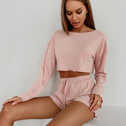 Frau in rosa Lounge-Set, bestehend aus langärmligem Crop-Top und Shorts, sitzt vor weißem Hintergrund. Bequeme Damenmode, Freizeitkleidung.