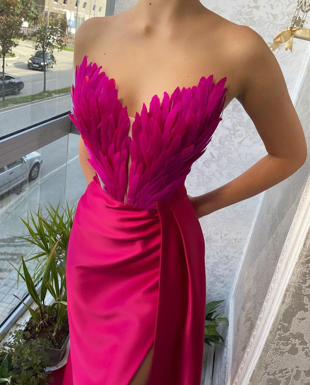 Frau in elegantem, pinkem Abendkleid mit Federmuster am Dekolleté, steht am Fenster. Luxuriöse Mode, Abendgarderobe, stilvolles Design.