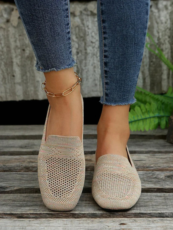 Damenfüße in hellbeigen, gestrickten Loafers auf Holzboden, kombiniert mit blauen Jeans. Modische, bequeme Sommerschuhe für Damen.