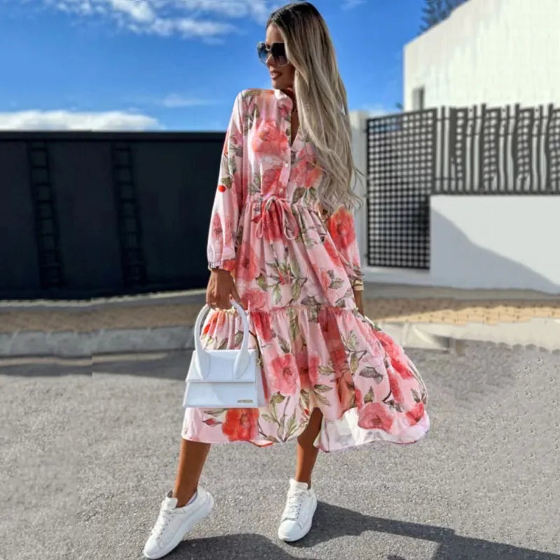 Frau in langem, geblümtem Kleid mit weißen Sneakers und Handtasche, steht vor modernem Gebäude. Sommermode, stilvoll, urbaner Look.