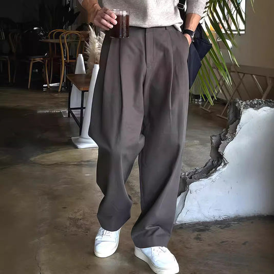 Mann in grauer, weiter Hose und weißen Sneakern hält ein Getränk in der Hand. Moderne Herrenmode, lässiger Stil, Café-Hintergrund.