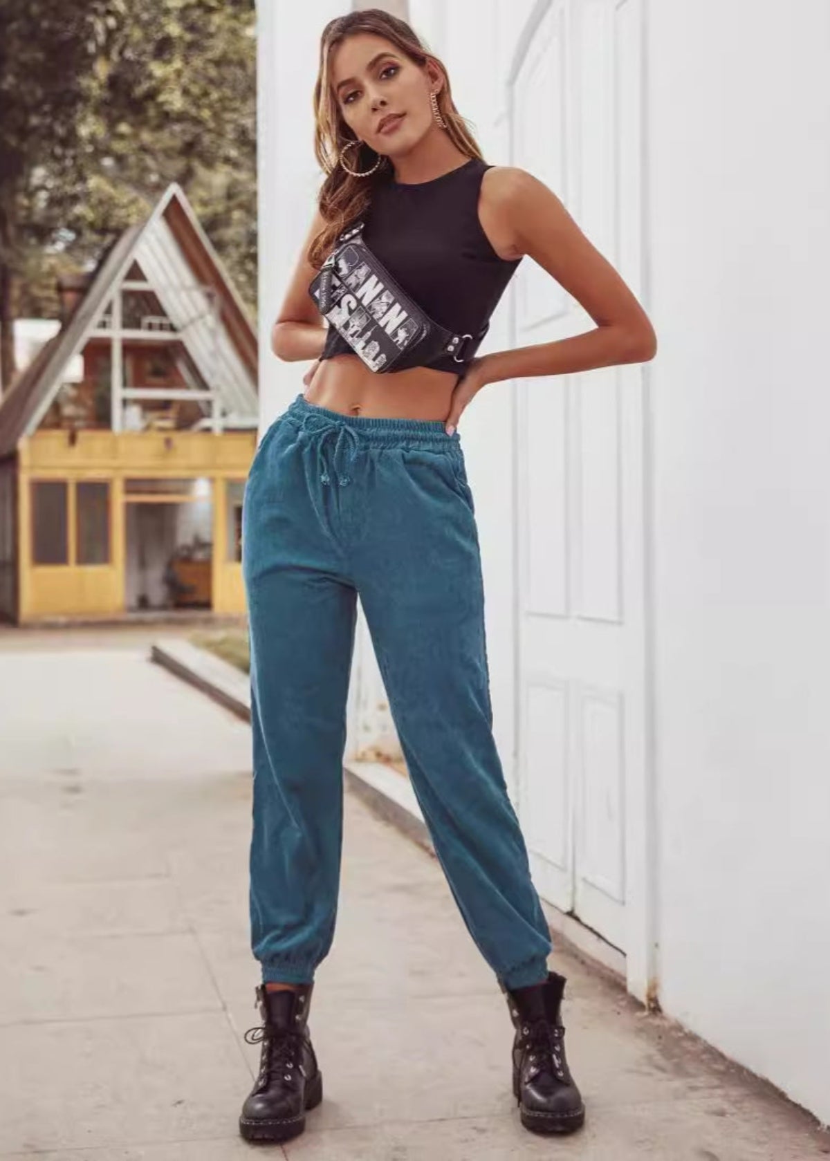 Frau in schwarzem Crop-Top und blauer Jogginghose, trägt schwarze Stiefel und Bauchtasche, steht vor einem modernen Gebäude im Freien.