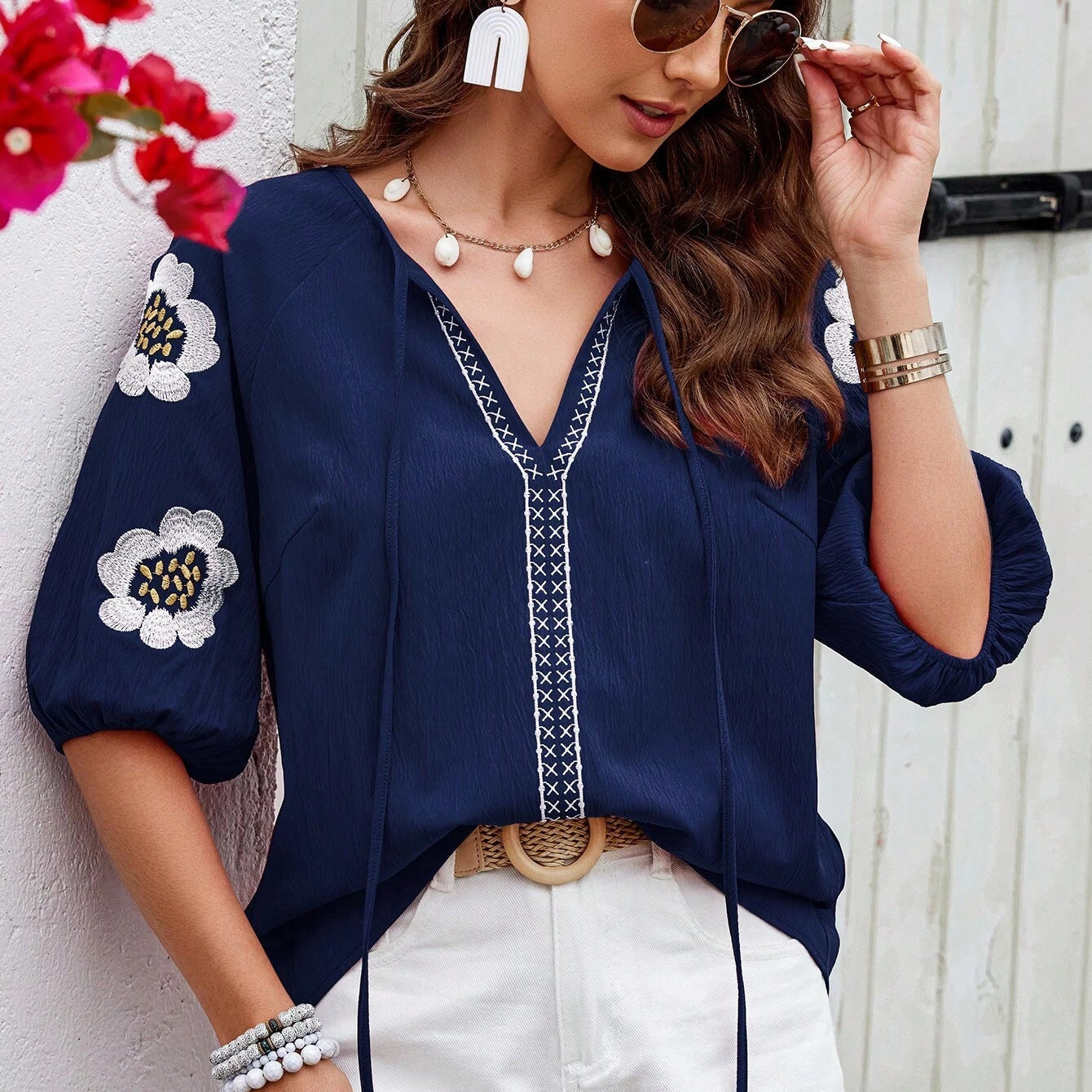 Frau in dunkelblauer Bluse mit Blumenstickerei, V-Ausschnitt und weißen Hosen, trägt Sonnenbrille und Schmuck, vor weißer Wand mit Blumen.