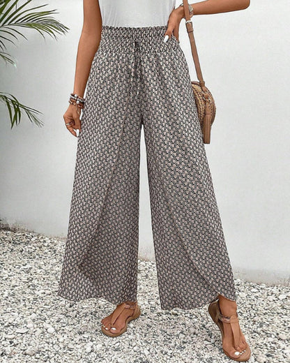 Frau in gemusterten, weiten Sommerhosen, Sandalen und weißem Top, steht auf Kiesweg. Modetrend, Damenmode, lässiger Sommerlook.