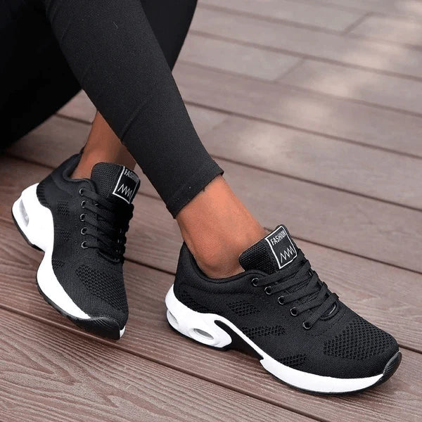 Schwarze Damen-Sneaker mit weißer Sohle, atmungsaktivem Mesh-Obermaterial und Schnürung, getragen auf Holzterrasse. Ideal für Sport und Freizeit.