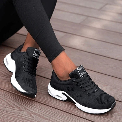 Schwarze Damen-Sneaker mit weißer Sohle, atmungsaktivem Mesh-Obermaterial und Schnürung, getragen auf Holzterrasse. Ideal für Sport und Freizeit.