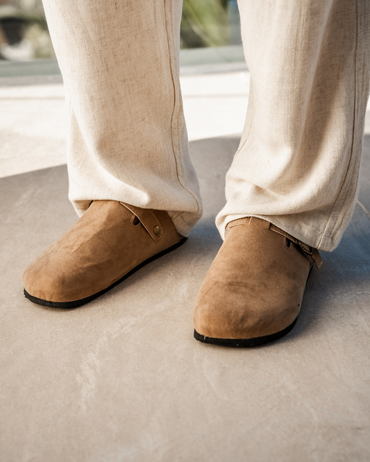 Beige Wildleder-Slipper auf grauem Boden, getragen mit hellen Leinenhosen. Modische, bequeme Schuhe für Herren. Ideal für Freizeit und Sommer.