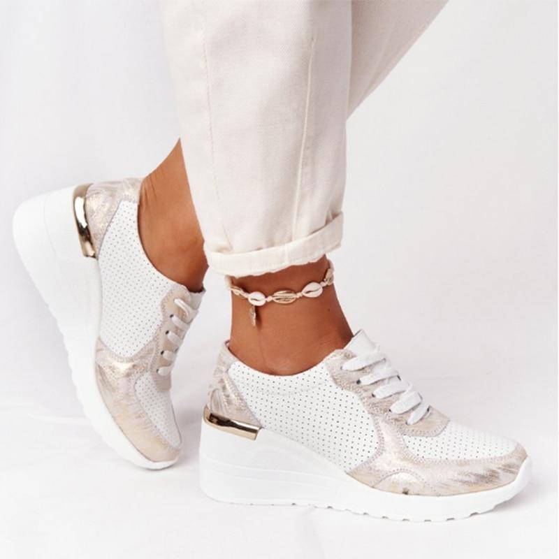 Weiße Damen-Sneaker mit Keilabsatz, goldenen Akzenten und Perforationen, getragen mit weißer Hose und Muschel-Fußkettchen. Modische Freizeitschuhe.
