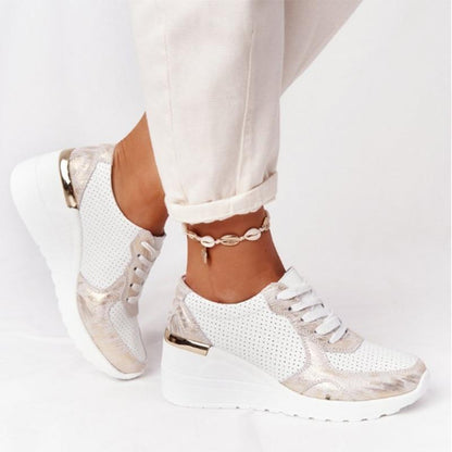 Weiße Damen-Sneaker mit Keilabsatz, goldenen Akzenten und Perforationen, getragen mit weißer Hose und Muschel-Fußkettchen. Modische Freizeitschuhe.