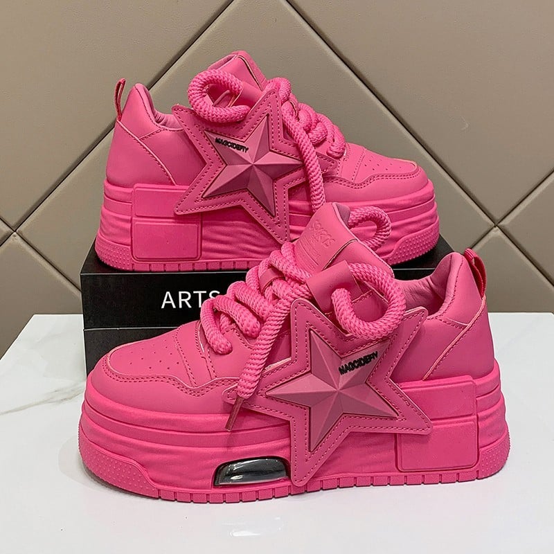 Pinkfarbene Plateau-Sneaker mit Stern-Design, dicken Schnürsenkeln und markantem Sohlenprofil auf schwarzem Karton vor beigem Hintergrund.