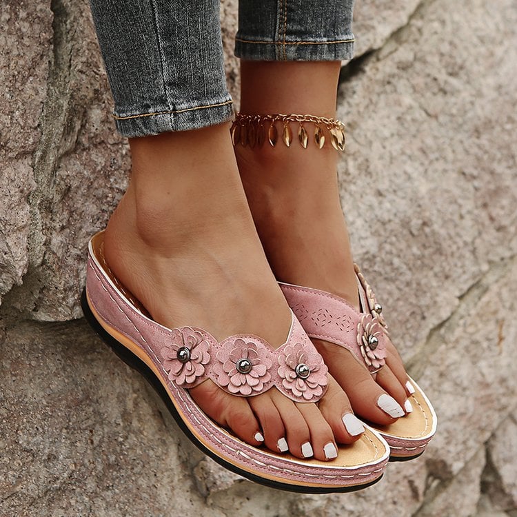 Damenfüße in rosa Sandalen mit Blumenmuster und Zehentrenner, Jeans, goldene Fußkette, auf Steinwand. Sommermode, bequeme Schuhe.