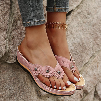 Damenfüße in rosa Sandalen mit Blumenmuster und Zehentrenner, Jeans, goldene Fußkette, auf Steinwand. Sommermode, bequeme Schuhe.