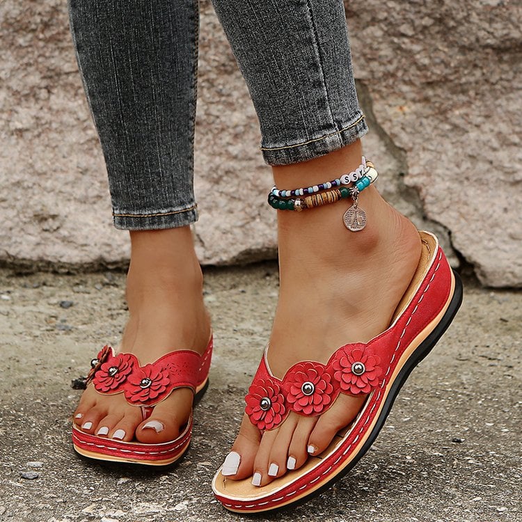 Frau trägt rote Sandalen mit Blumenmuster, Jeans und Perlenfußkettchen. Modische Sommerschuhe, Damenmode, bequeme Sandalen.