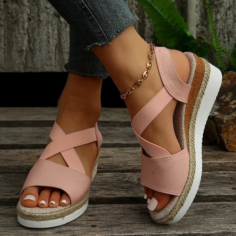 Damenfüße in rosa Sandalen mit Keilabsatz und geflochtener Sohle, kombiniert mit Jeans und goldener Fußkette, auf Holzboden.