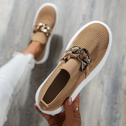 Beige Damen-Sneaker mit Strick-Obermaterial und goldener Kettendekoration, getragen von Person in weißen Jeans auf Holzfußboden.