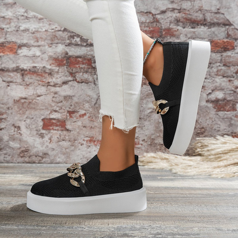 Schwarze Damen-Sneaker mit Plateau und goldener Kettendekoration, getragen zu weißen Jeans vor einer Ziegelwand. Modische Freizeitschuhe.