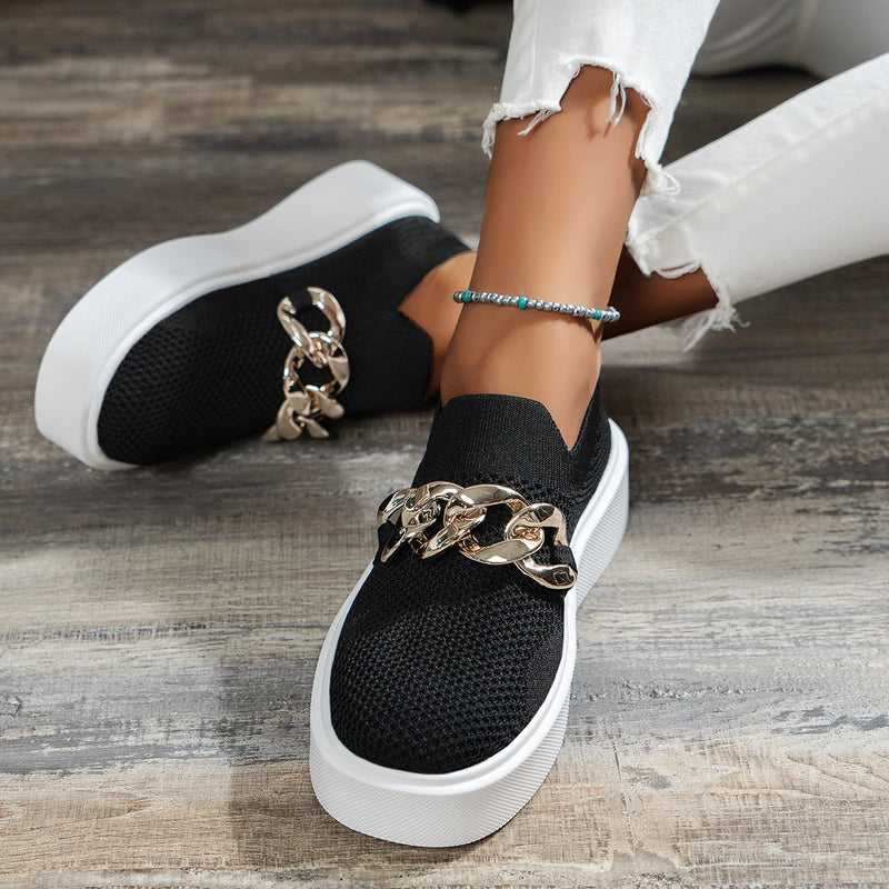 Schwarze Slip-On-Sneaker mit goldener Kette, weiße Sohle, getragen von Person in zerrissenen weißen Jeans. Modische Damen-Schuhe, trendiger Look.