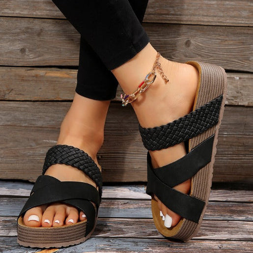 Schwarze geflochtene Sandalen mit Keilabsatz an Frauenfüßen, kombiniert mit schwarzer Hose und goldfarbenem Fußkettchen auf Holzboden.