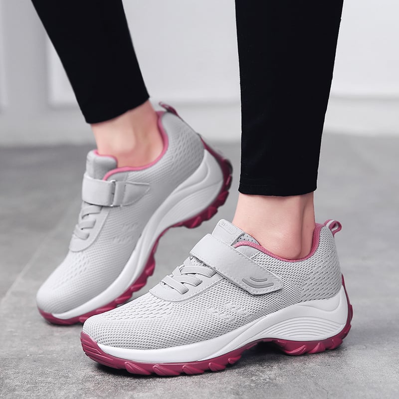 Graue Damen-Sneaker mit Klettverschluss und rosa Sohle, getragen auf grauem Boden. Bequeme Sportschuhe für Alltag und Fitness.
