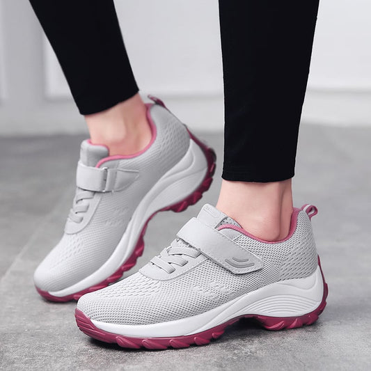 Graue Damen-Sneaker mit Klettverschluss und rosa Sohle, getragen auf grauem Boden. Bequeme Sportschuhe für Alltag und Fitness.
