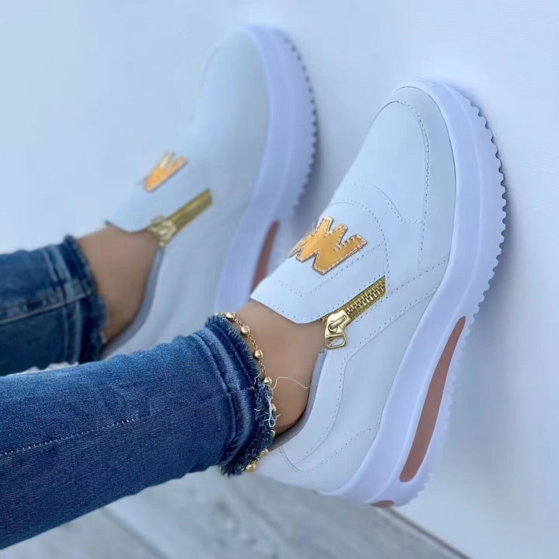 Weiße Damen-Sneaker mit goldenen Reißverschlüssen und Initialen, getragen mit blauen Jeans. Modische Freizeitschuhe, ideal für trendige Outfits.
