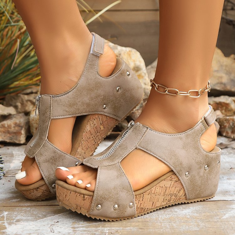 Beige Keilsandalen mit Reißverschluss und Nieten, Korksohle, getragen von einer Person auf Holzboden. Modische Sommerschuhe für Damen.