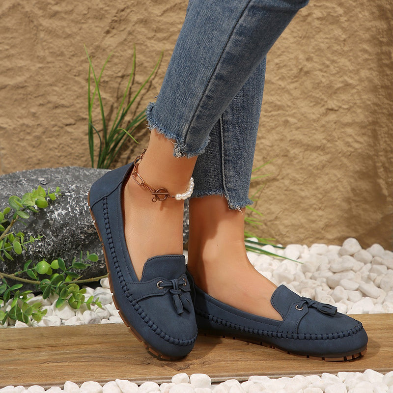 Damenfüße in blauen Mokassins auf Kieselsteinen, Jeans hochgekrempelt, stilvoller Freizeitlook, Outdoor-Szene, modische Schuhe, bequeme Passform.
