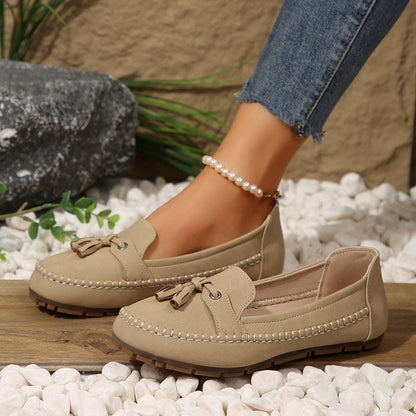 Beige Mokassins mit Quasten, getragen von einer Person mit Jeans und Perlenknöchelband, auf weißen Kieselsteinen. Elegante Damen-Schuhe, Freizeitlook.