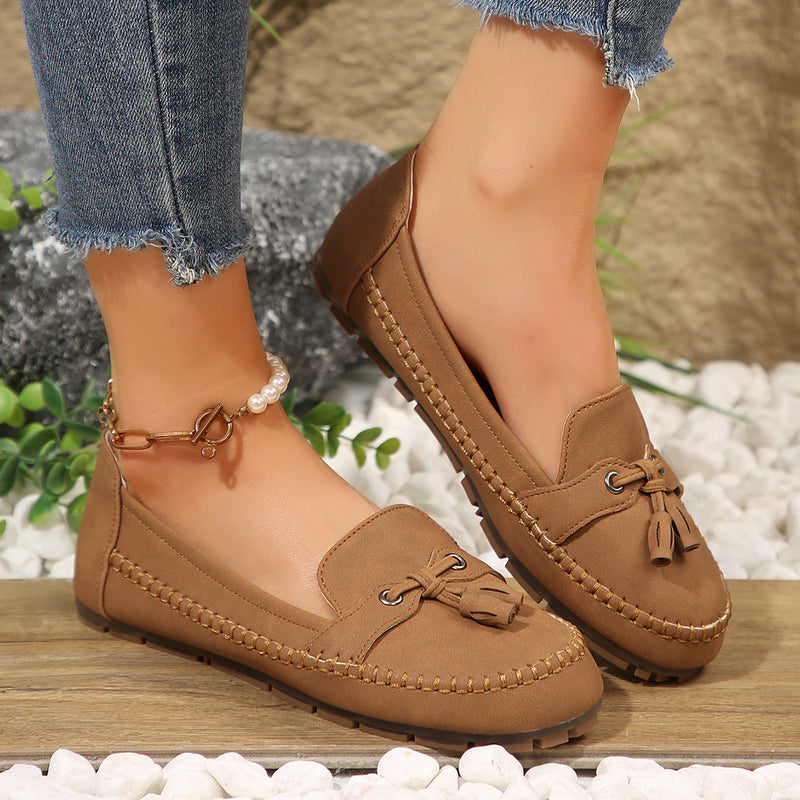 Braune Mokassins mit Quasten, getragen von einer Person in Jeans, auf Kieselsteinen. Modische Damen-Schuhe, bequem und stilvoll.