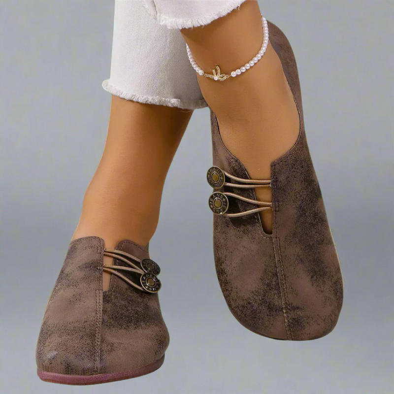 Braune Damen-Loafers mit dekorativen Knöpfen, getragen mit weißer Jeans und Perlenfußkettchen. Modische, bequeme Schuhe für den Alltag.